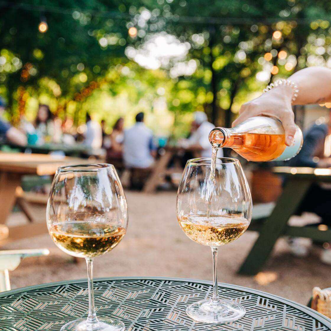 Wine tasting event setup at Cork and Screw Wine Garden with two glasses being filled, outdoor setting, string lights.