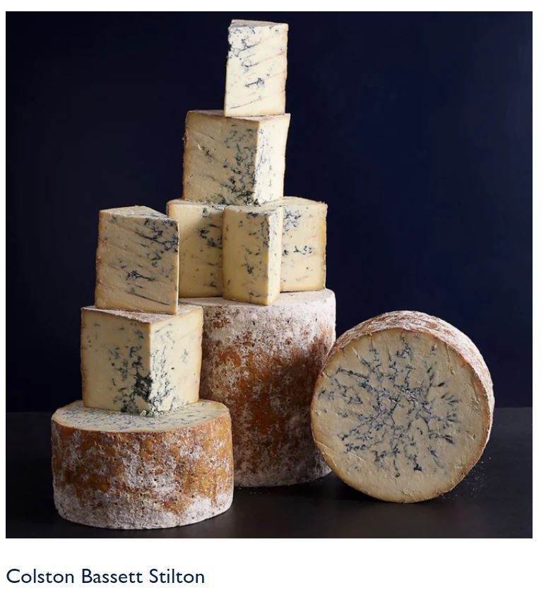 Colston Bassett Stilton cheese wheels and wedges stacked on a dark background.